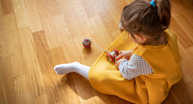 criança sentada no chão de madeira, rodando um pião de brinquedo no piso